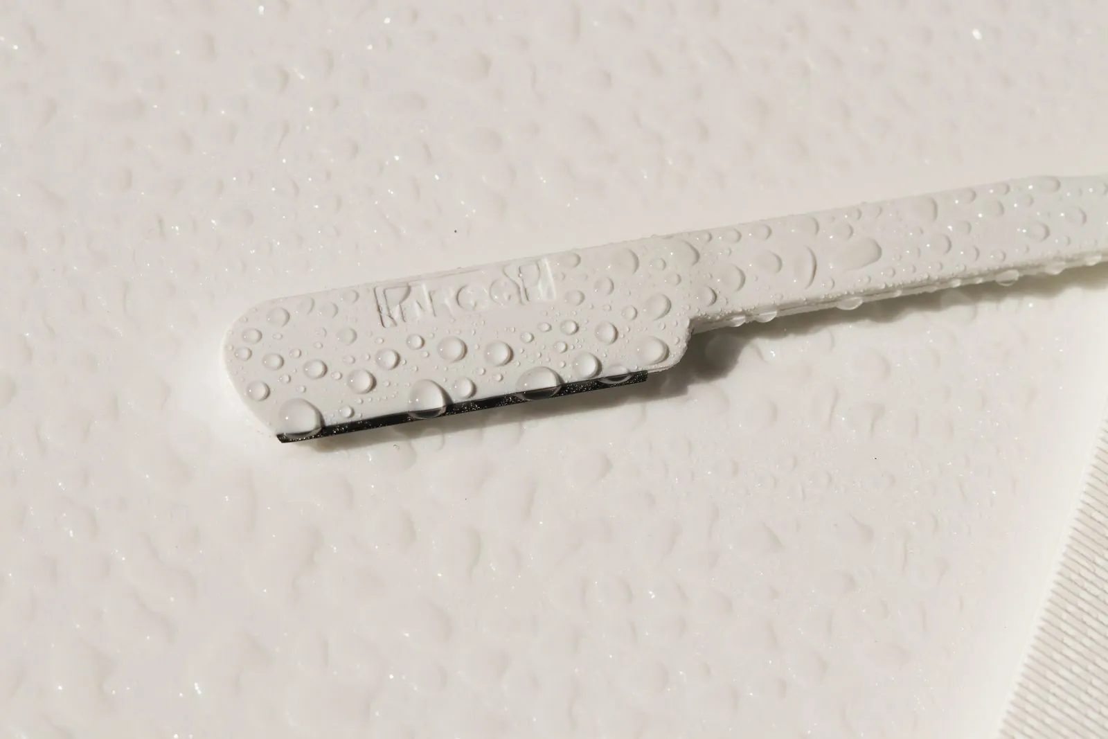 A white single-blade dermaplaning razor with a short straight edge, lying on a wet white surface