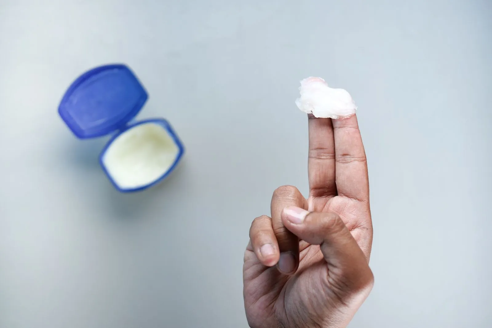Two fingertips holding a dollop of petroleum jelly next to an open tub