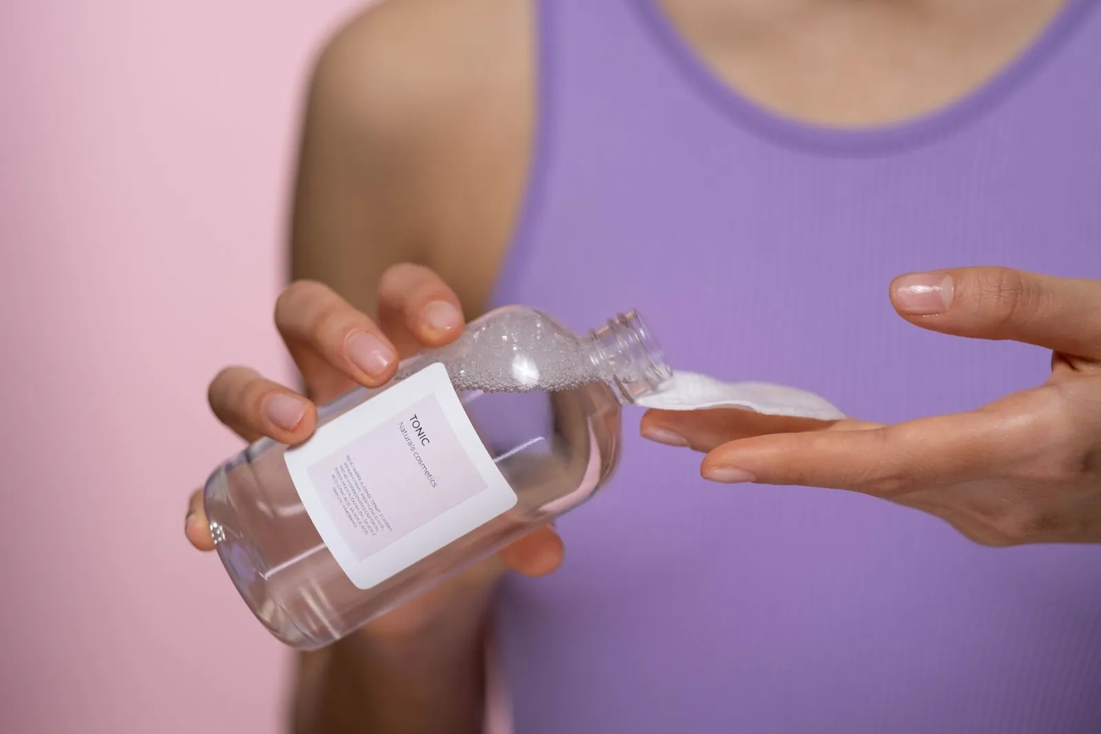 Hands pouring clear liquid toner from a bottle onto a cotton pad