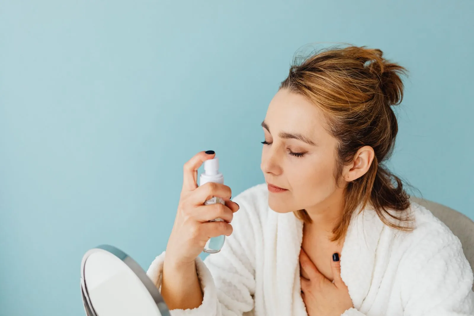 Woman in a bathrobe misting a spray toner onto her face in front of a small mirror