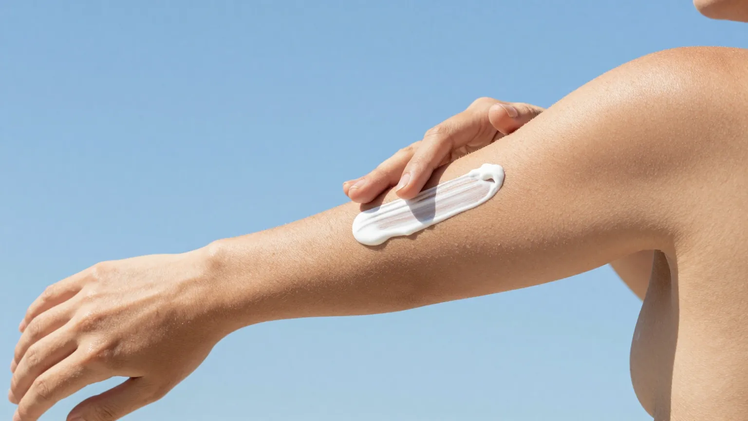 Woman smoothing a streak of white sunscreen along her forearm under a clear blue sky